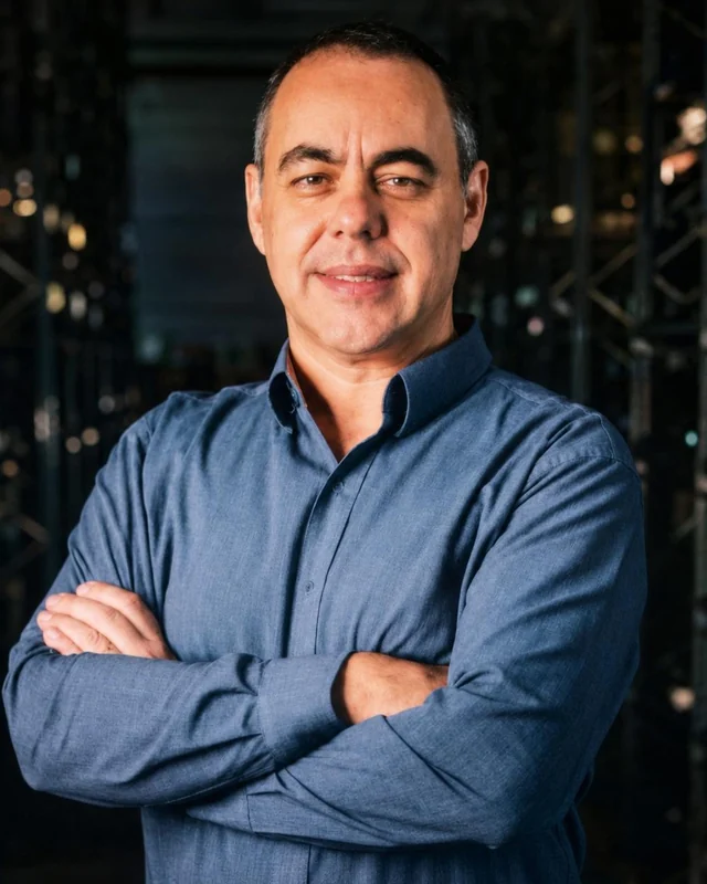 Miguel Rossato, de camisa azul e braços cruzados, em frente a estantes de um galpão